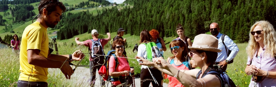 Luca Stefenelli tende uno spago ad una delle persone del gruppo che lo segue. Tutti si trovano fermi su una strada asfaltata n montagna. Sullo sfondo si vede una distesa di abeti.