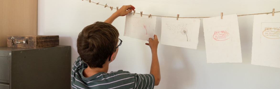 Bambino intento ad appendere, con delle mollette, delle stampe ad un filo. Queste stampe sono su fogli di carta bianca formato A4