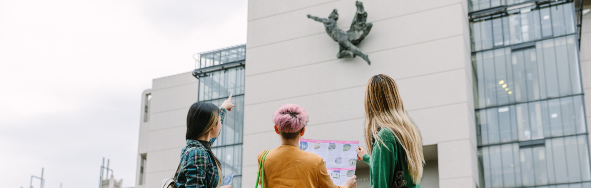 Three people looking at a map stood in front of a concrete building and pointing up at a sculpture on the wall.