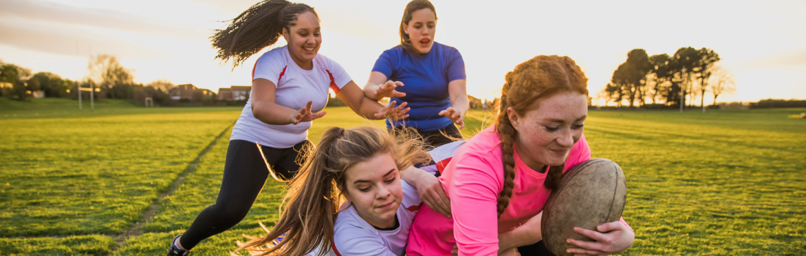 Four girls play rugby on a grassy field at sunset. One carries the ball while others move to tackle her, showing teamwork and determination.
