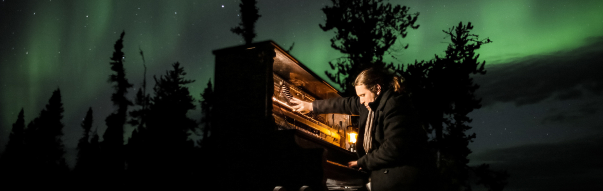 A man sits at a grand piano, his hands are blurry due to movement, above him is an immersive view of the auroura bo