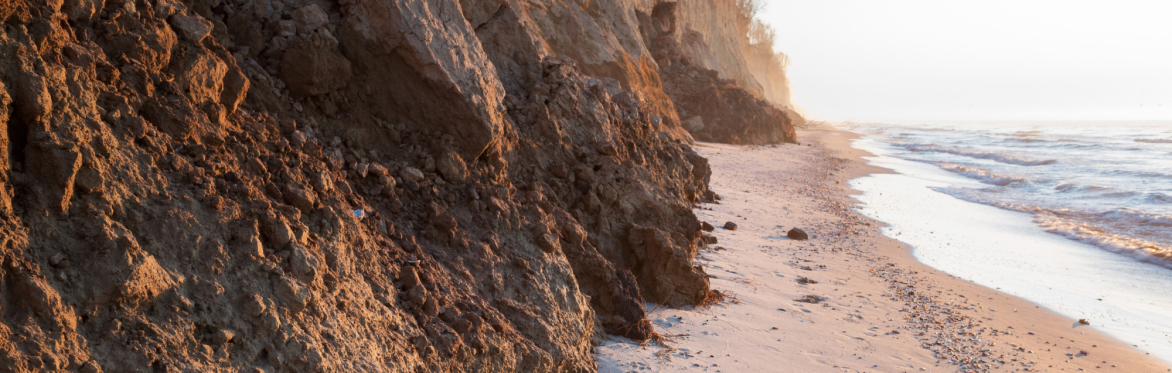 Curved rock barrier extends into calm sea under a blue sky, creating a serene, peaceful coastal scene. Cooper Education logo appears on the top right hand side.