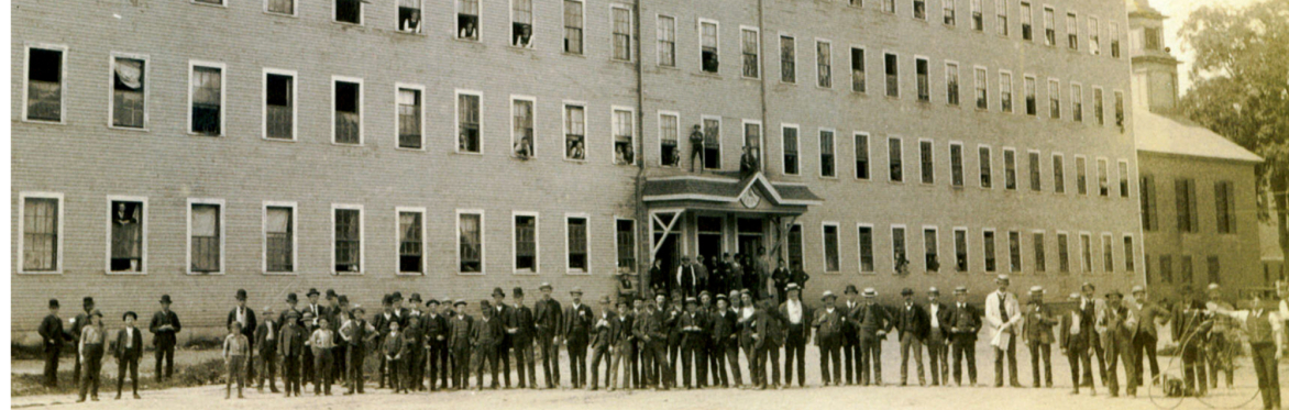 Black and white photo of a large wooden factory building with a line of men standing if front of it posing for the camera.