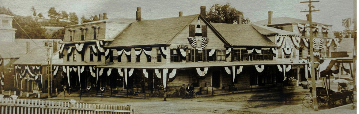 Black and white photo showing the corner of a village street with two and three story buildings festooned in patriotic bunting.