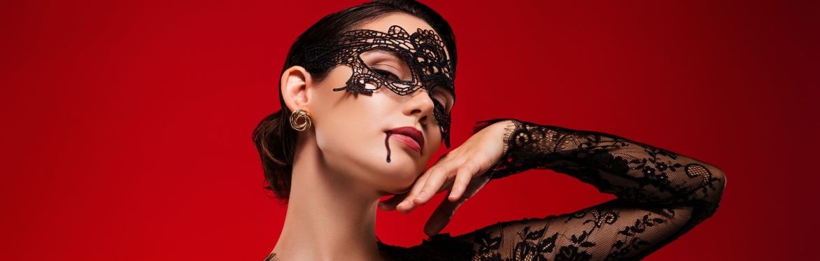 Woman in an ornate black lace masquerade mask and black dress against a deep red background, with a trickle of blood at the corner of her mouth.