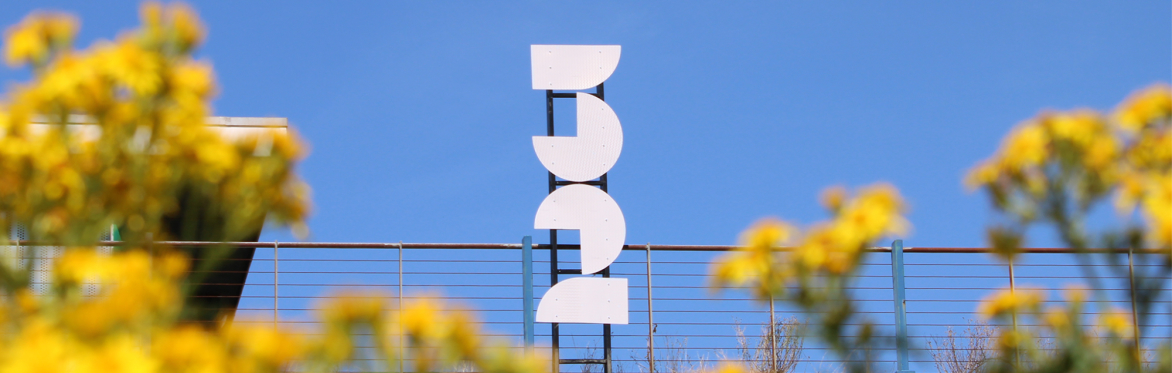 A large white sign read 3 2 (the No.32) strapped to a balcony with the blue sky in the background and yellow flowers in the foreground.