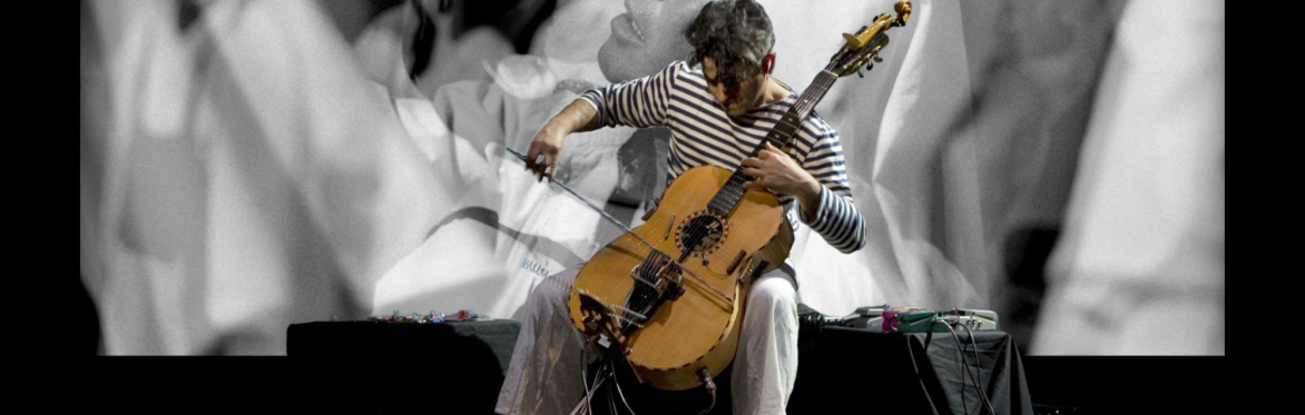 Paolo Angeli plays his prepared Sardinian guitar on stage, with a large black-and-white projection of two faces behind him.