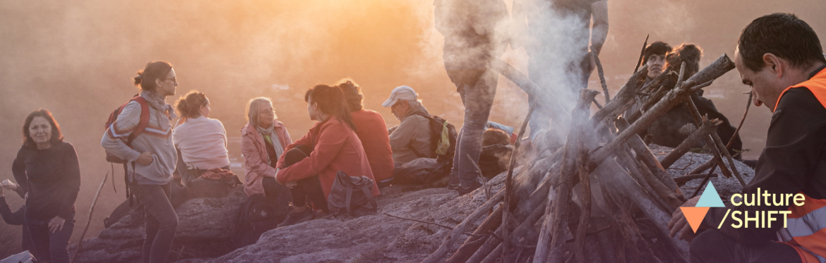People sat on a mountain peak at sunset. In the foreground, someone lights a fire.