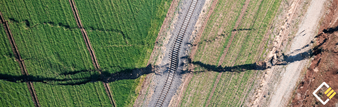 Aerial view of green fields with a dark crack running through them, crossing a railroad track. The image conveys tension and disruption.
