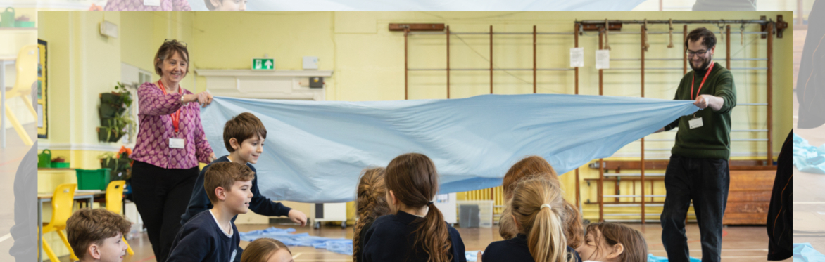 Children and facilitators using blue fabric to represent a river during a creative storytelling and drama workshop in a school hall.