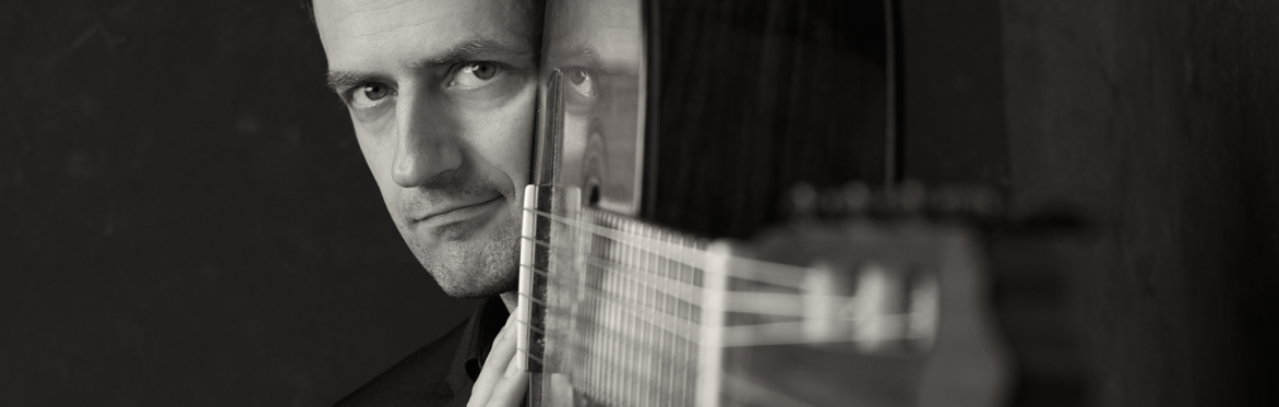 Black-and-white portrait of a male classical guitarist in a dark suit, holding his guitar vertically.