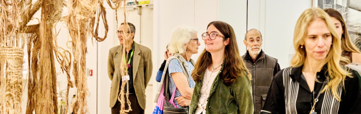 People gathered in a white walled gallery looking at the artworks. In the centre is a person with glasses wearing a green jacket looking up at a textile sculpture.