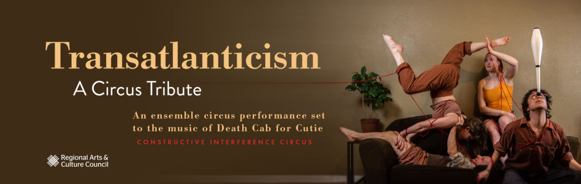 Acrobats dressed in earthy colors hold each other up and balance a white juggling club on their face while sitting on a couch.