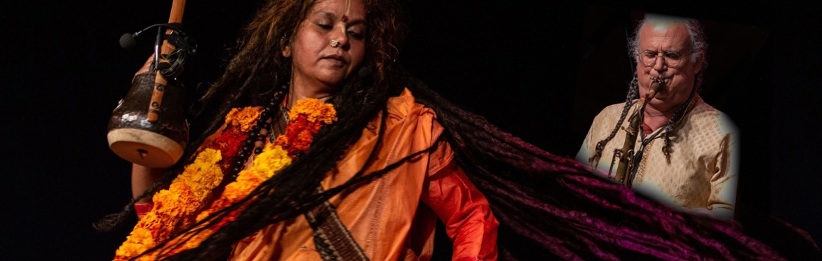 Parvathy Baul spinning, eyes shut, holding an ektara,dreadlocks splayed out to the side, behind stands a man playing saxophone.