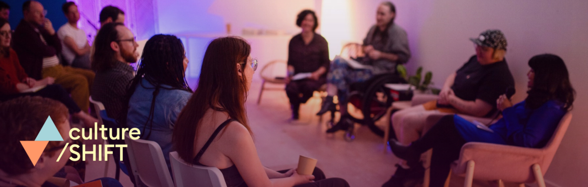 A group of people sitting in a softly lit room listening to a panel of speakers.