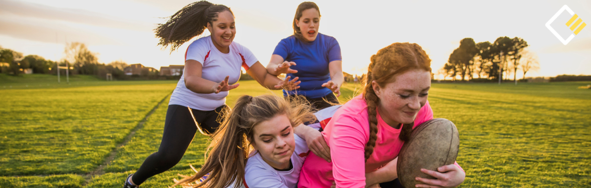 Four girls play rugby on a grassy field at sunset. One carries the ball while others move to tackle her, showing teamwork and determination.