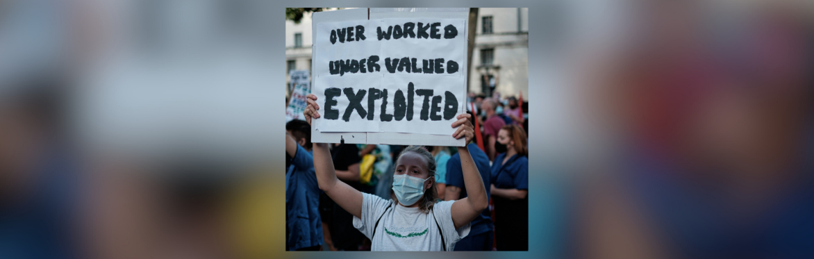 A protestor holding up a sign which reads 'Over worked, under valued, exploited.'