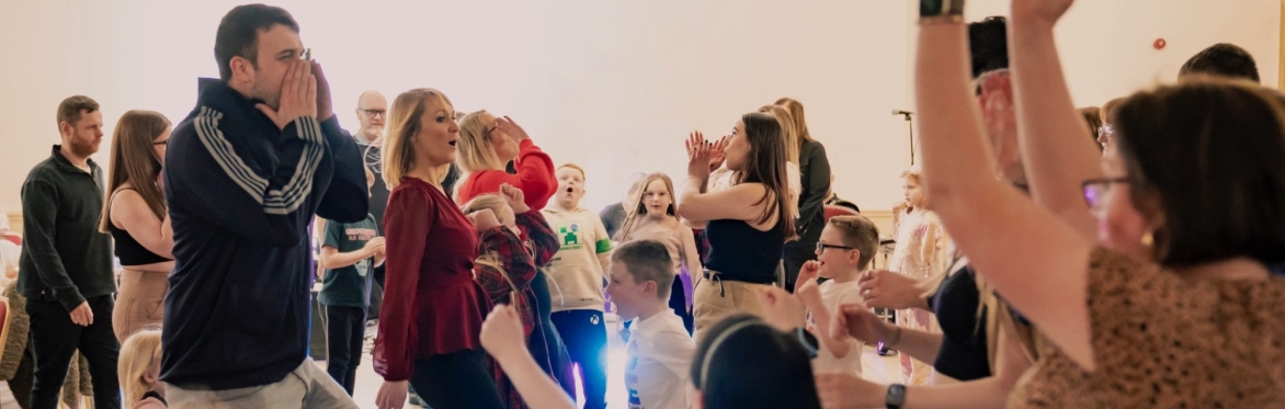 A lively group of adults and children enjoying a ceilidh dance in a bright hall, full of energy and joy.