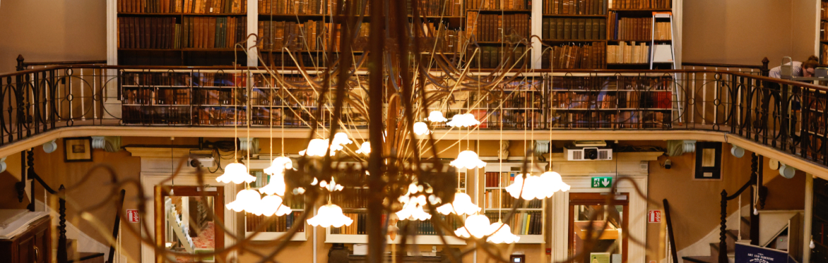 Behind a grand 19th century chandelier, with glass light fixtures, a balcony and rail is visible, with dark wooden bookshelves stacked with leatherbound books.