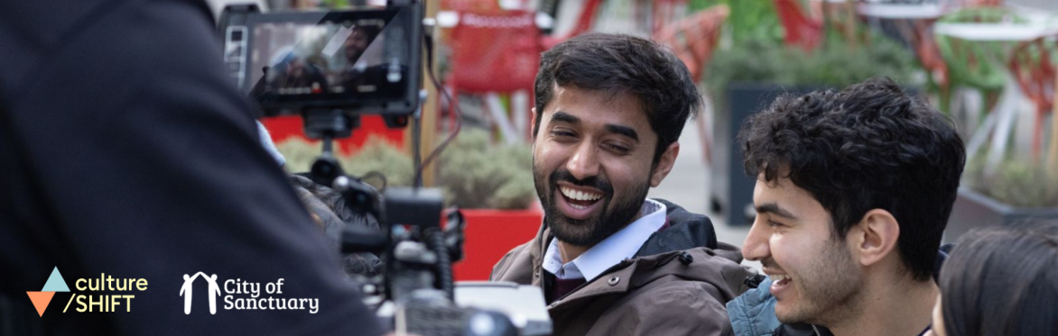 Two men laughing while being filmed by a digital camera.