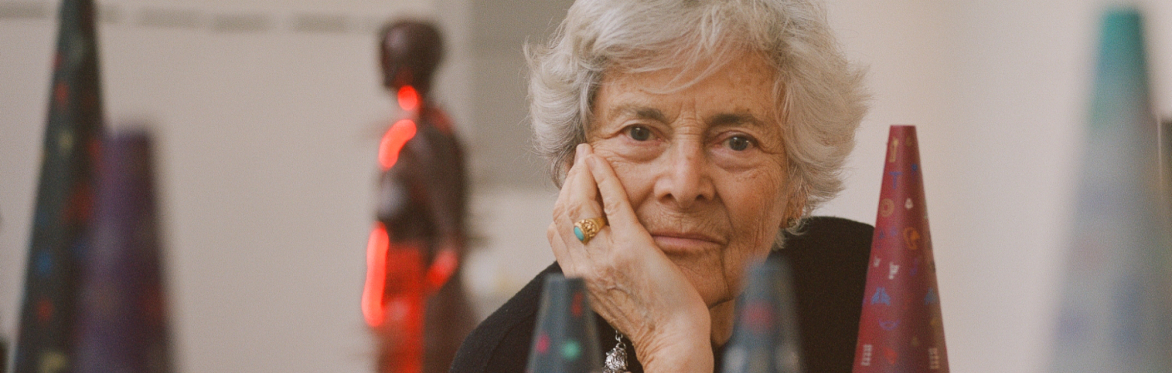 A woman with short grey hair looks at the camera with her head resting on her hand. Around her are cone shaped sculptures, some in and some out of focus.