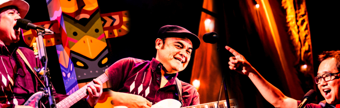 Three musicians performing on stage; the central figure plays guitar and smiles under warm stage lighting, flanked by a singer at a microphone on the left and another performer on the right, with colo