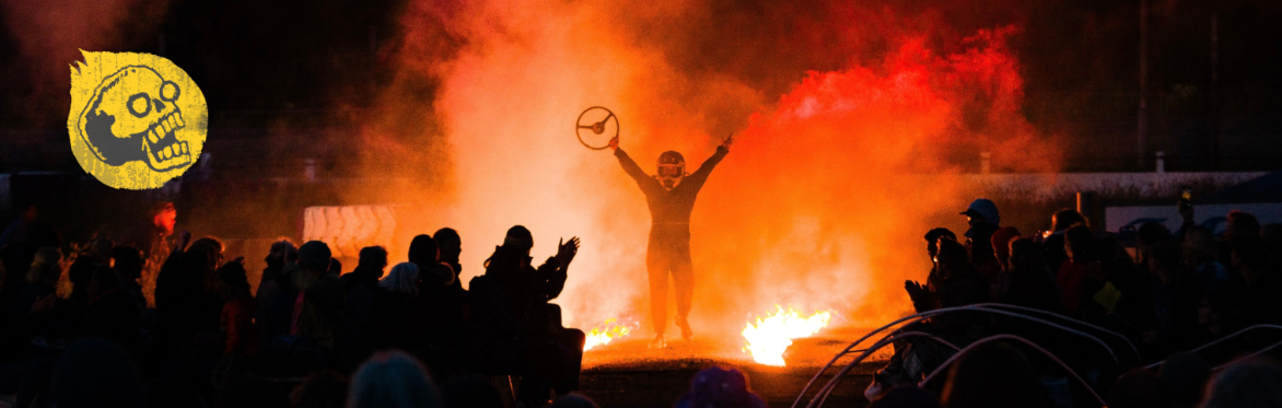 Maddy emerges through the flames and smoke holding a steering wheel. The audience are clapping in the foreground