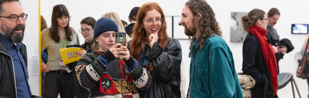 A group of people stood surrounded by art in a white walled gallery. They are smiling and one is taking photos on a mobile phone.