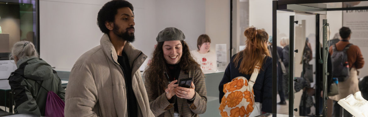 People stood in a museum surrounded by objects in glass cases. In the centre are two people stood together whilst one smiles at their phone.