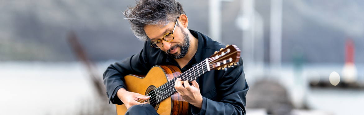 Pablo Garibay, wearing glasses with grey hair, plays a classical guitar while seated on a wooden dock.