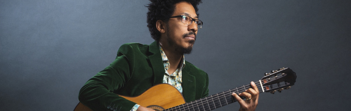 A man with dark, curly hair and glasses, wearing a green velvet blazer and patterned shirt, plays a classical guitar.