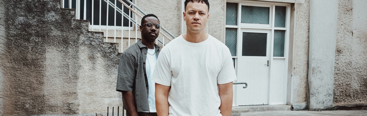 Image of the two band members of Tekoa standing in front of grunge building with hands in pockets.