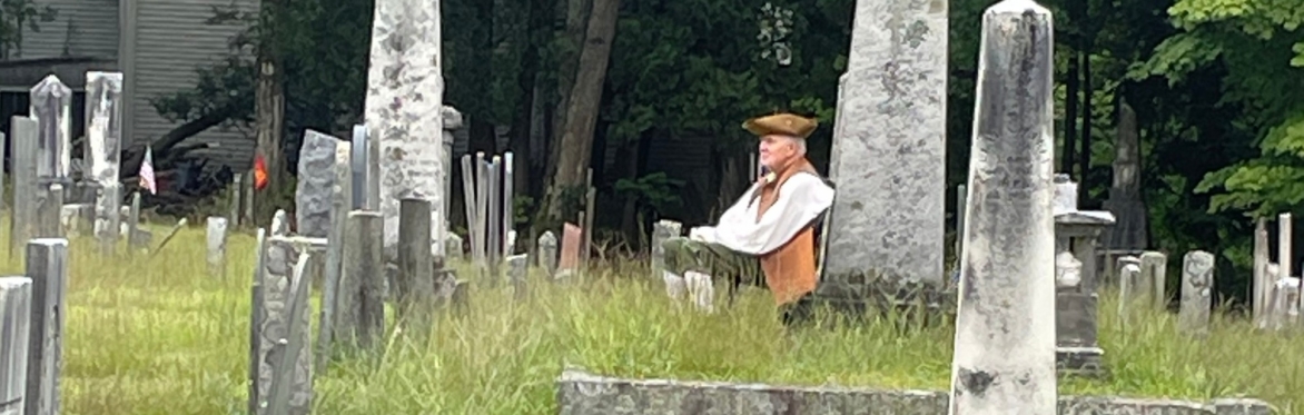 Man in a tricorn hat, vest and breeches seated at the base of a gravestone gazing into the distance.