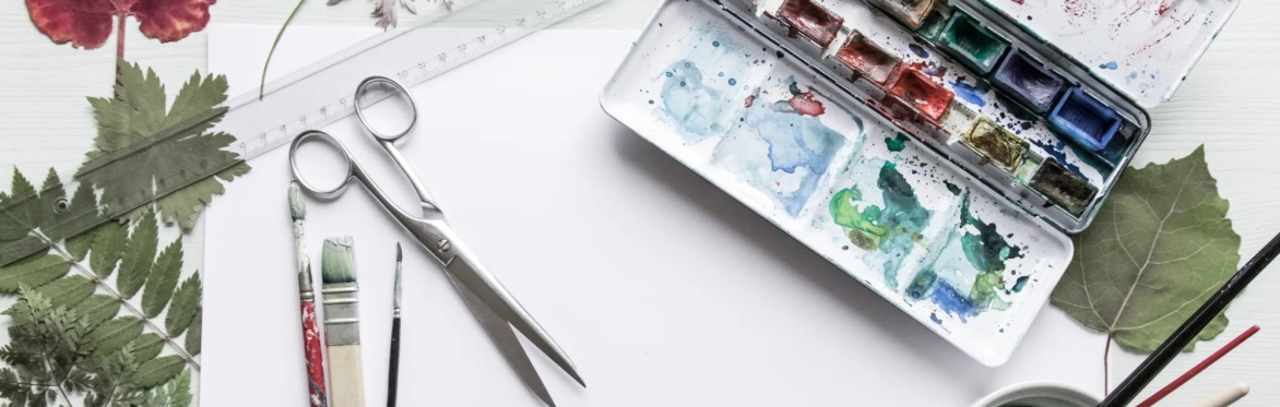 Artistic flat lay composition with a white background, featuring a used watercolor palette with remnants of blue and red paints, a pair of metal scissors, a transparent ruler, two paintbrushes with st