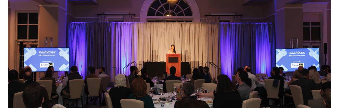 a view from the back of the banquet room of Myra Ruiz welcoming attendees of the 2024 InnoVision awards. Myra is standing on a stage behind a podium. Attendees are seated at round tables.