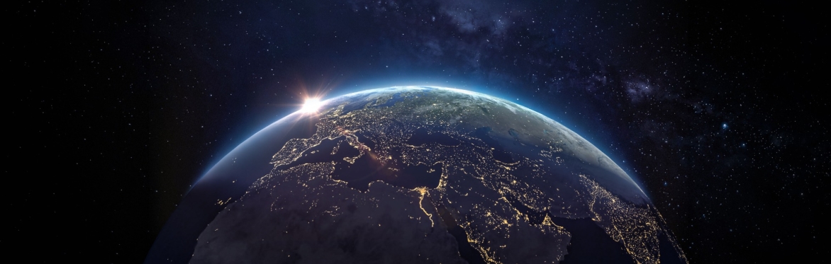 View of Earth from space during nighttime, showing illuminated city lights across Europe, the Middle East, and parts of Africa and Asia. The Sun is peeking over the horizon, creating a warm glow again