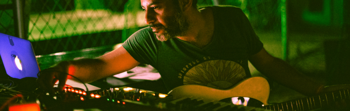 Maurice Louca holds an electric guitar while operating a synthesizer.