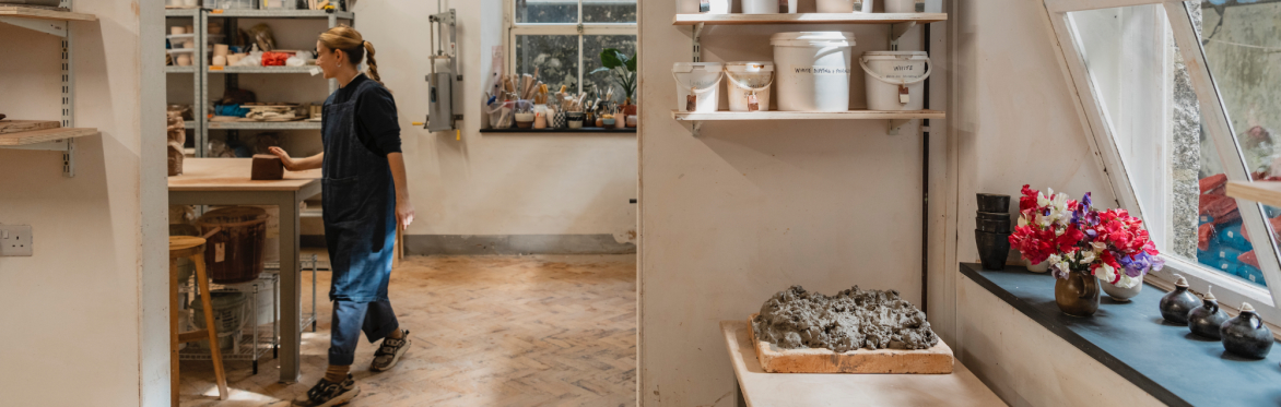 View of the inside of a ceramics studio showing a person next to a table near shelves of ceramics kit as well as fired and unfired clay