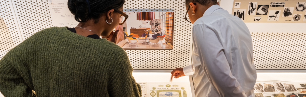 A tour guide talks to a guest admiring printed works, furniture parts, and collected ephemera in a vitrine.