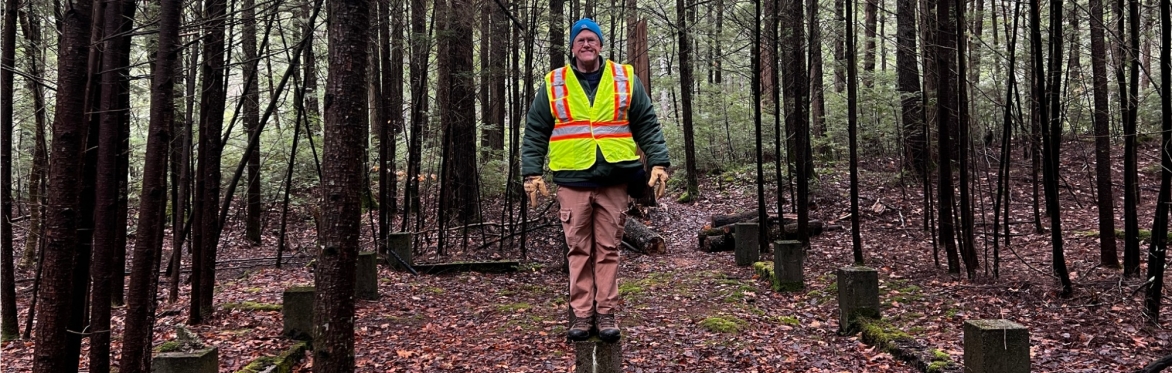 A man in a safety vest stands perched on two pieces of wood stuck in the ground in the woods. The wood appears to be part of a an old foundation or structure.