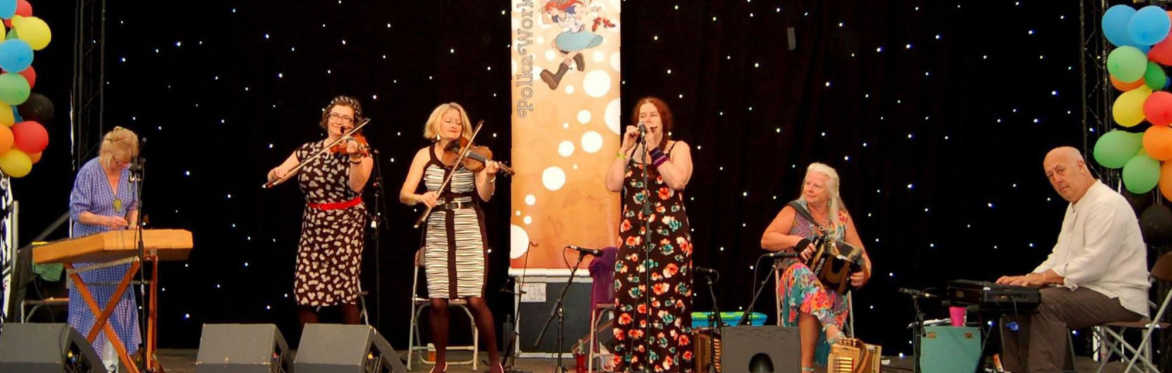 7 members of the band: 5 sitting in a row, 2 standing behind. Some are holding instruments - a saxophone and a diatonic accordian. Behind them are a selection of paintings on the walls.