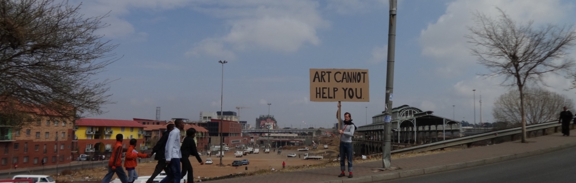 A white man holding a large cardboard sign that reads "Art Cannot Help You" on the side of a street in a non-western/non-European environment.