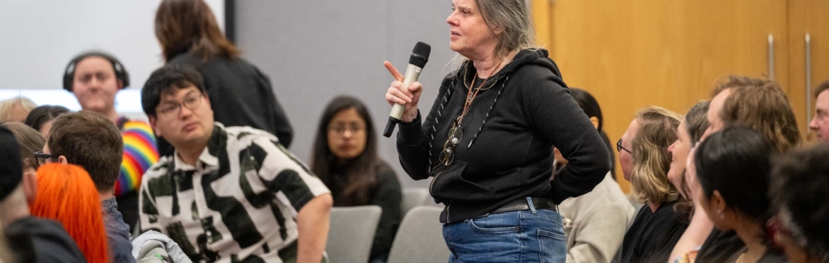 Image shows a women speaking into a microphone. She is in a large room with other people watching her speak.