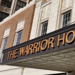 Historic hotel entrance with a black marquee reading “THE WARRIOR HOTEL” on a cream stone and brick building.