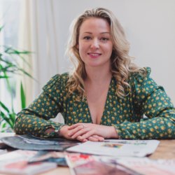 picture of Susanne Bearne, a white woman in a green dress, sat at a desk with newspapers in front of her
