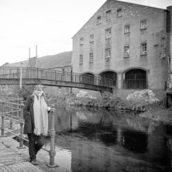 A woman stands buy a river, near an old bridge.