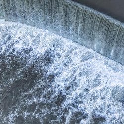 Aerial view of a curved dam with water cascading over, creating white foam.