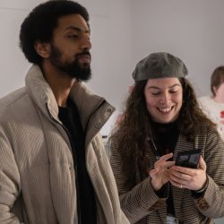People stood in a museum surrounded by objects in glass cases. In the centre are two people stood together whilst one smiles at their phone.