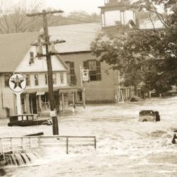 black and white photo showing a heavily flooded street with 1930s era cars in the water. The water is above the height of the tires.
