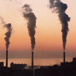 Factory smokestacks emit thick smoke against a colourful sunset sky, depicting pollution and industrial impact on the environment.